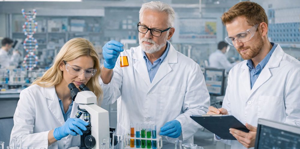 Drei Naturwissenschaftler in Laborkitteln arbeiten mit Mikroskop und Reagenzgläsern.